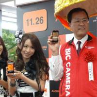 Raising the smart phone bar: KDDI Corp. Senior Vice President Takashi Tanaka pitches the new IS03 smart phone with members of the idol group SDN48 Friday at the Bic Camera electronics outlet in Tokyo\'s Yurakucho district. | KAZUAKI NAGATA PHOTO